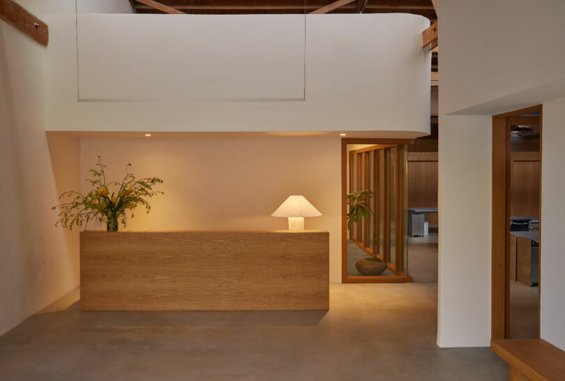 A minimalist reception area features a wooden desk with a lamp and a floral arrangement. The space has soft lighting, neutral walls, and modern decor.