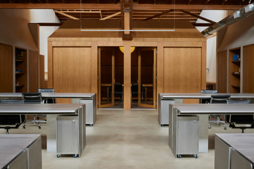 Modern office space with minimalist design featuring metal desks, ergonomic chairs, and a wooden central structure. The area has exposed beams and an industrial decor.