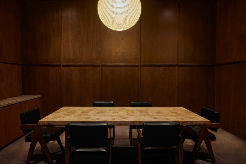 A dimly lit conference room features a rectangular wooden table surrounded by six black chairs, illuminated by a single large spherical pendant light. The walls are paneled in dark wood.