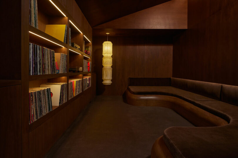 A dimly lit room with wooden walls and shelves filled with vinyl records. A curved cushioned seating area and a hanging cylindrical lamp add to the cozy ambiance.