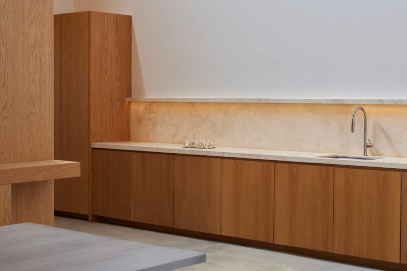 A minimalist kitchen with light wood cabinets, beige countertops, and a built-in sink beneath a simple wall shelf.