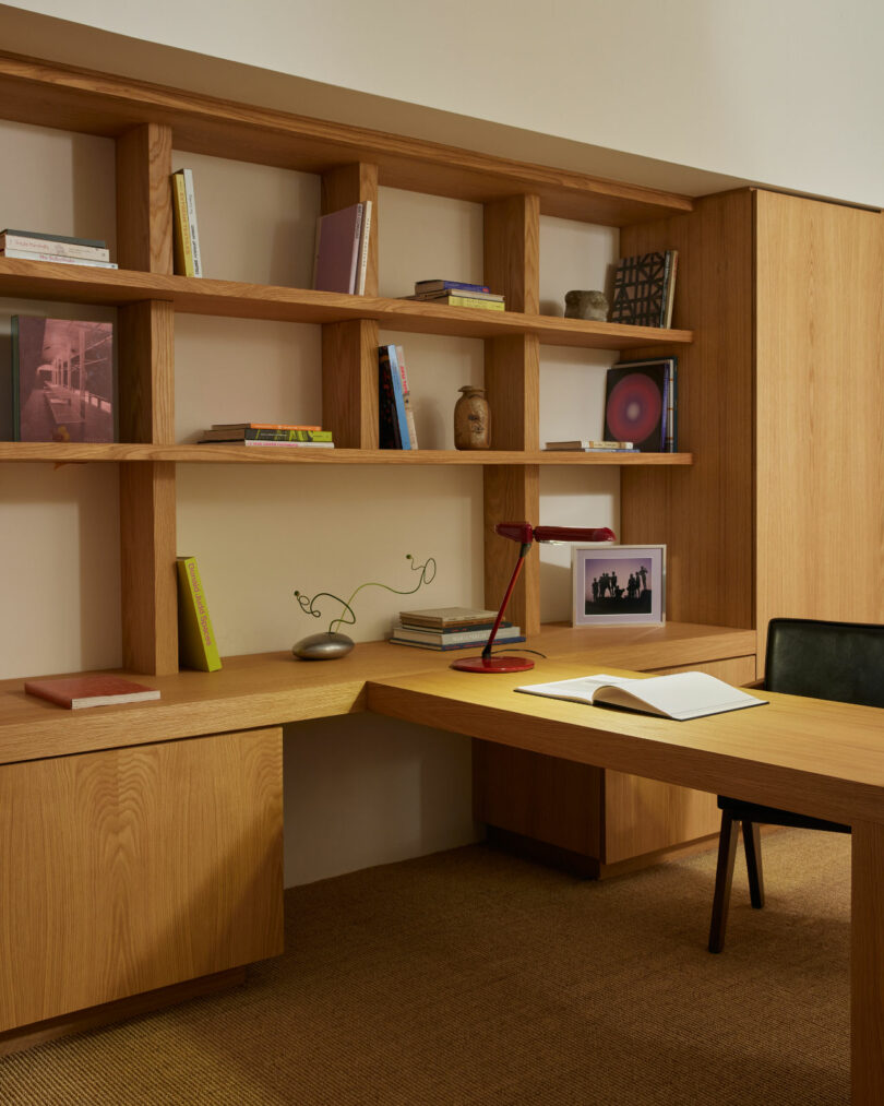 A modern home office with wooden desk and shelves, containing books, a red desk lamp, an open notebook, and artworks. A black chair is placed at the desk. The room has a carpeted floor.