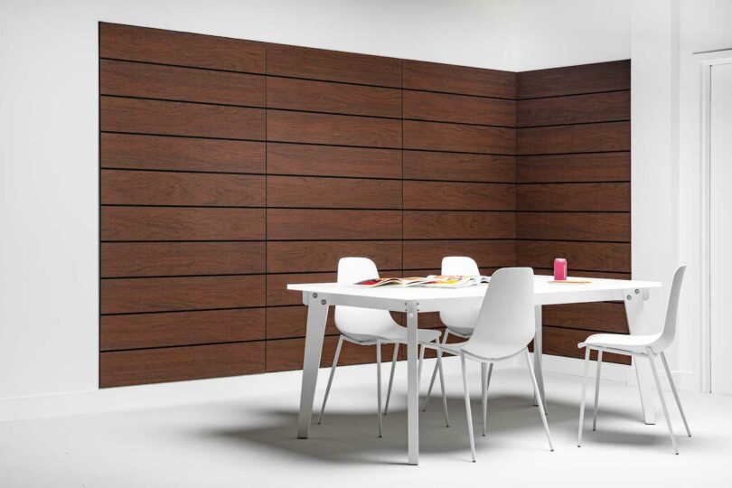 A modern minimalist room featuring a white table with four matching chairs. The walls are adorned with horizontal wood-like felt panels, and various items are placed on the table