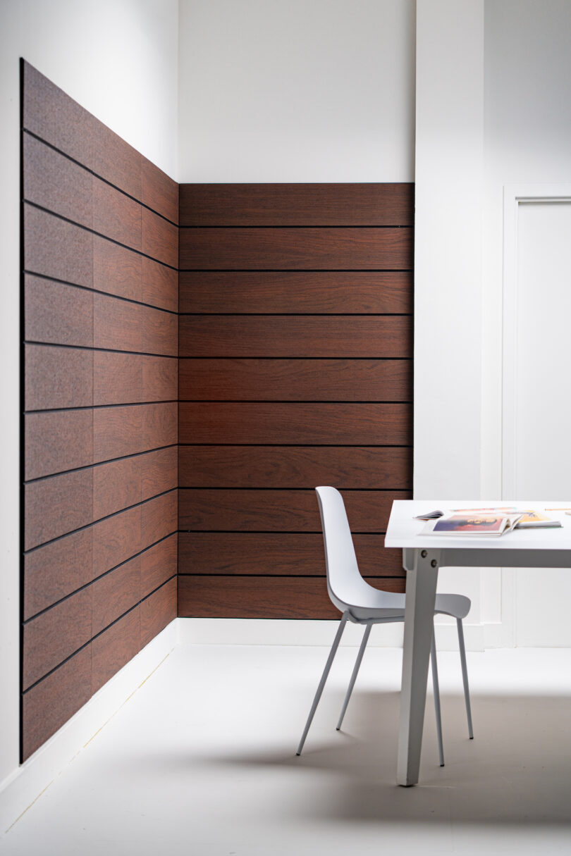 A minimalistic room features a white table with an open book and a white chair. The walls are partially covered with horizontal wooden panels. The space is well-lit with natural light