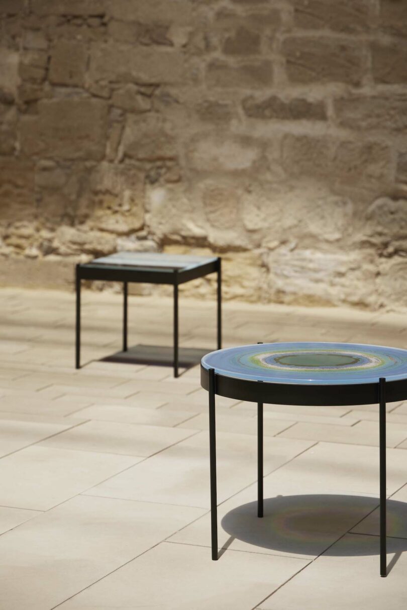 Two modern, circular tables with artistic, concentric designs on their tops are placed on a tiled floor against a stone wall background