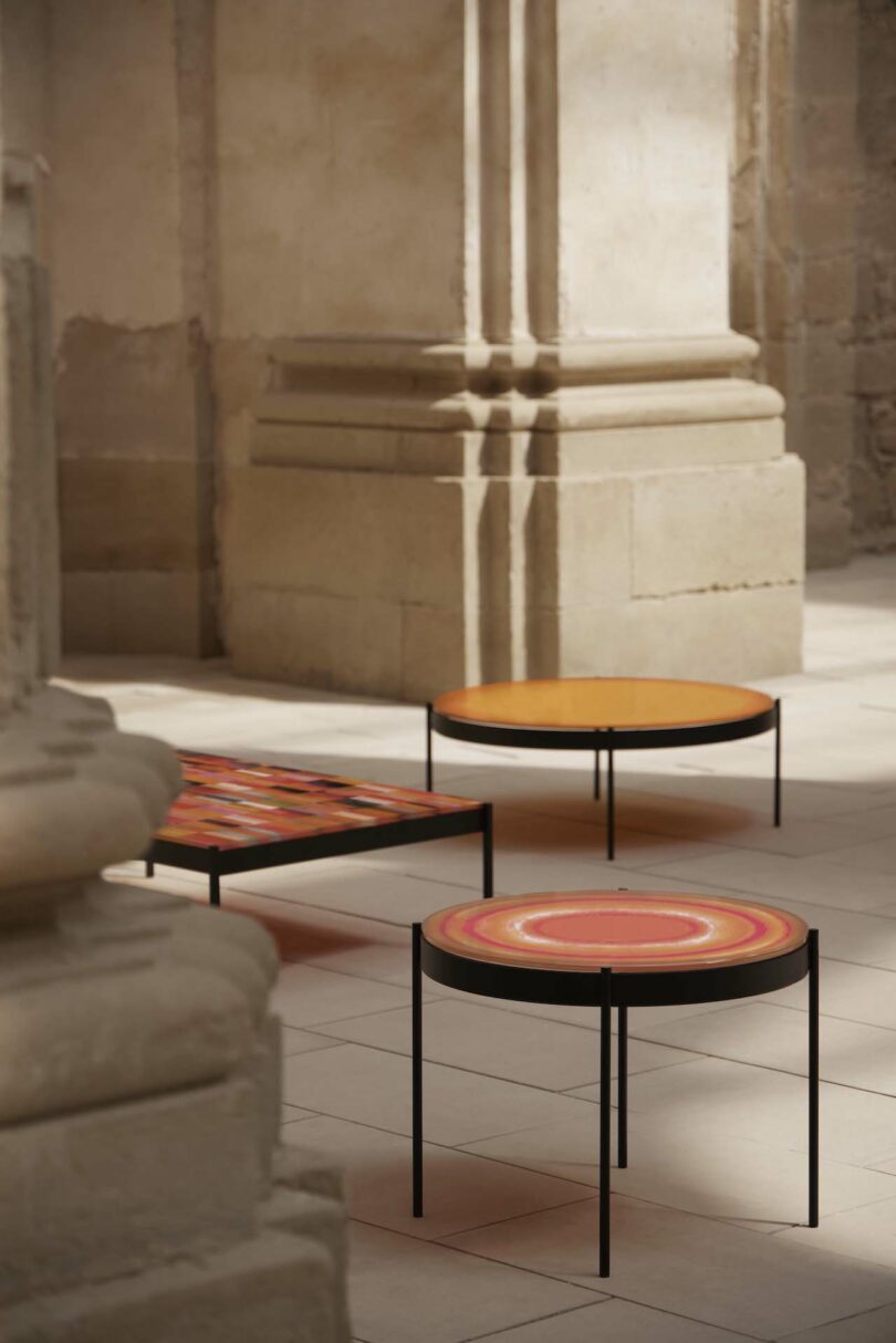 Three modern round tables with colorful tops and thin black legs are placed in a sunlit stone room with ornate architectural details