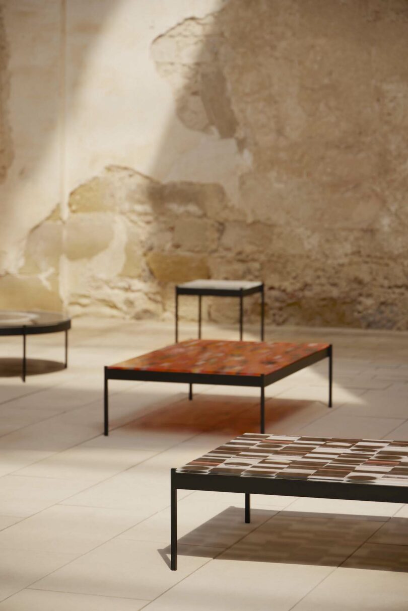 Four minimalist tables with thin metal legs and mosaic tops are arranged on a tiled floor against a textured, aged wall. Light is streaming in from the left