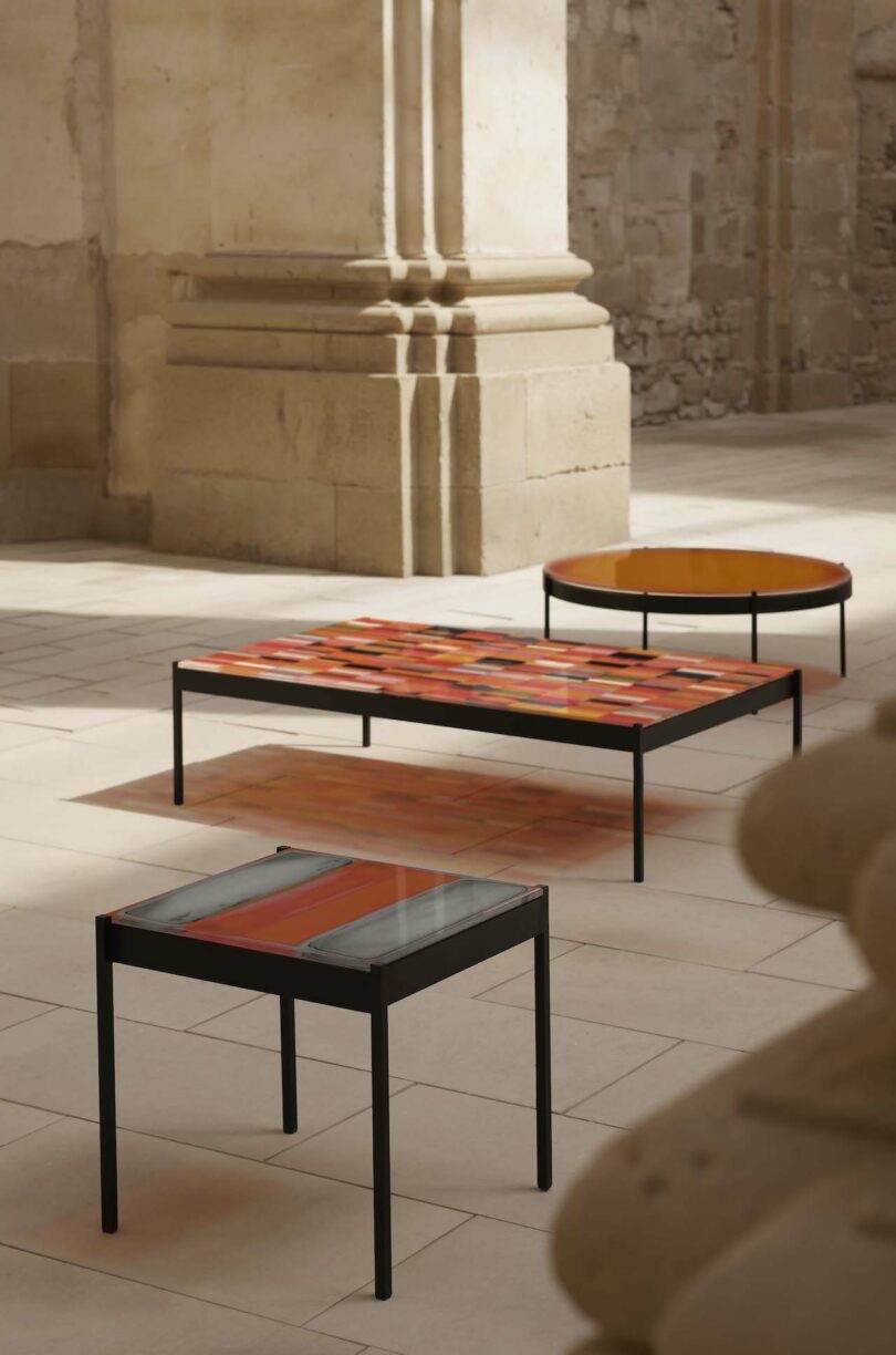 Three modern tables with vibrant orange and red surfaces stand in a stone courtyard with large columns and historic architecture