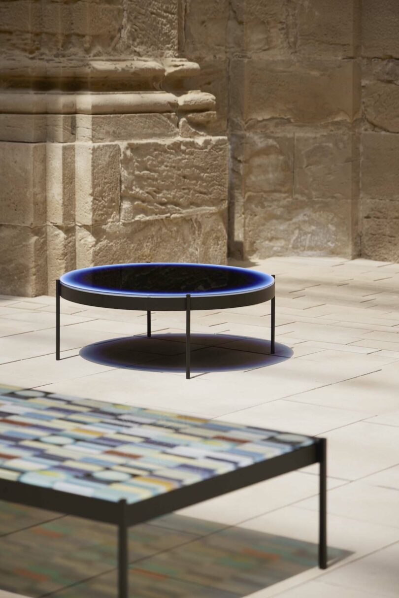 Contemporary round table with a blue edge and rectangular glass-top table in a sunlit stone courtyard.