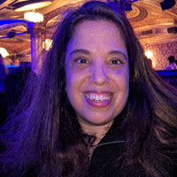 A woman with long brown hair is smiling while standing indoors with ornate lighting and decor in the background.