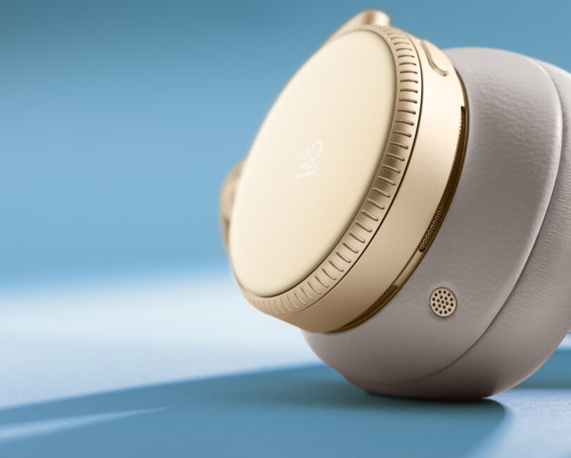 Close-up of Beoplay H100 gold and white over-ear headphones with a focus on the ear cup and surrounding details against a blue background.