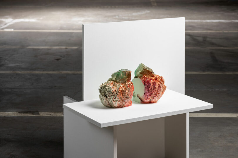 A sculptural piece with red, white, and green textures sits on a white table against a dark background, resembling organic rock formations.