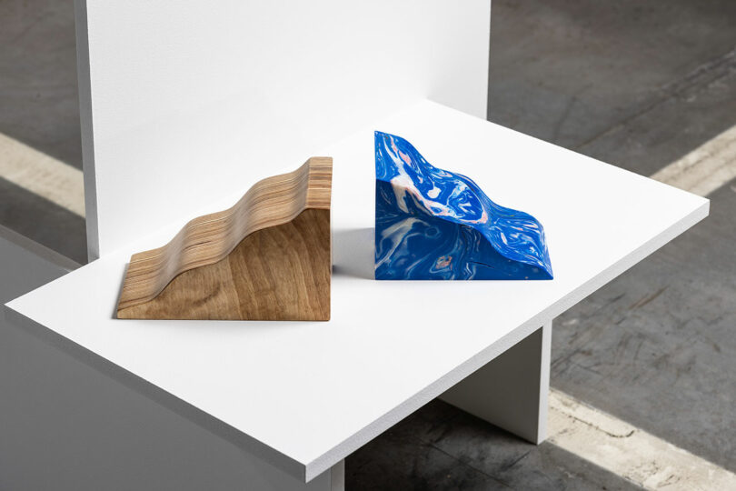 Two wave-shaped sculptures on a white surface: one is made of wood, and the other is blue with a glossy finish.