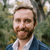 A man with short hair and a beard smiles at the camera. He is wearing a white shirt and a denim jacket, standing outdoors with green foliage in the background.