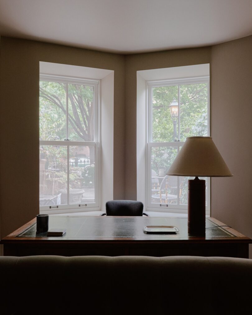 A simple office workspace features a desk with a lamp, two trays, a cup, and faces two windows displaying a view of trees and an outdoor lamp post.