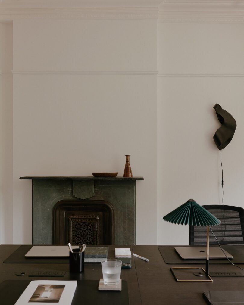 A minimalist workspace with two desks, a green lamp, a laptop, a notebook, and a cup on each. A decorative wall piece and an old fireplace are visible in the background.