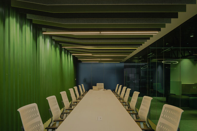 A modern conference room with a long table and white chairs, green walls, and a geometric ceiling design.