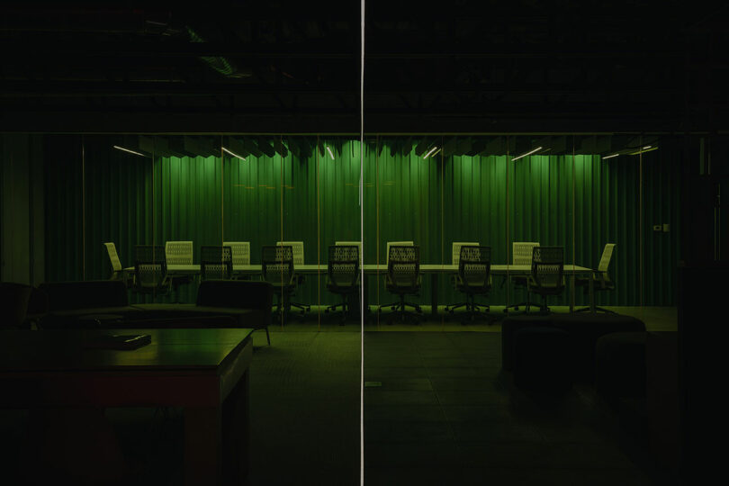 A dimly lit conference room with a long table and chairs against a green, vertically paneled wall.