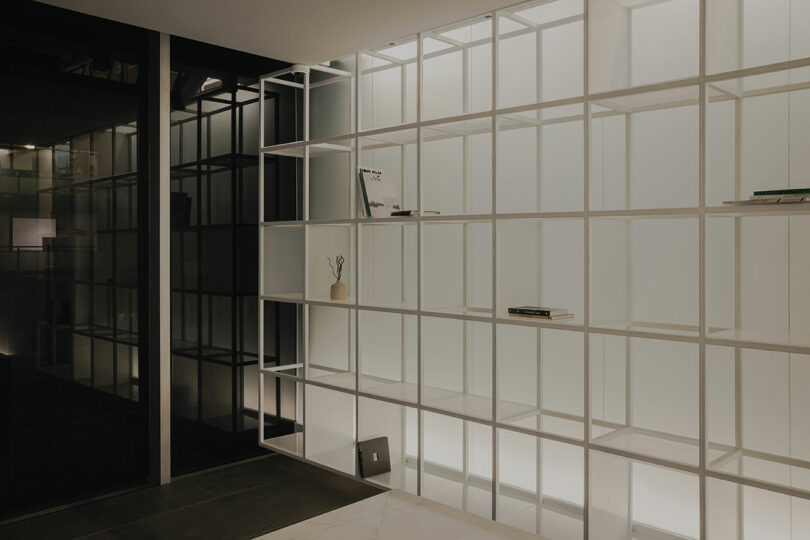 Minimalist room with a large white grid shelf against a backlit wall. Sparse items, including a vase and books, are placed on the shelves. Dark flooring and a reflective glass wall are visible.