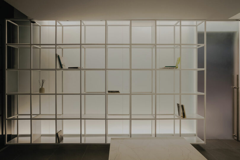 A minimalist room features a backlit, empty white shelving unit with a few scattered books and decorative items.