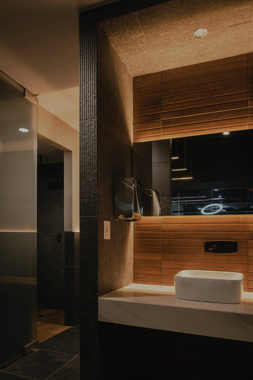 Modern bathroom with warm lighting, featuring a white rectangular sink on a marble countertop, a large mirror with wooden paneling, and a partially visible glass shower enclosure.