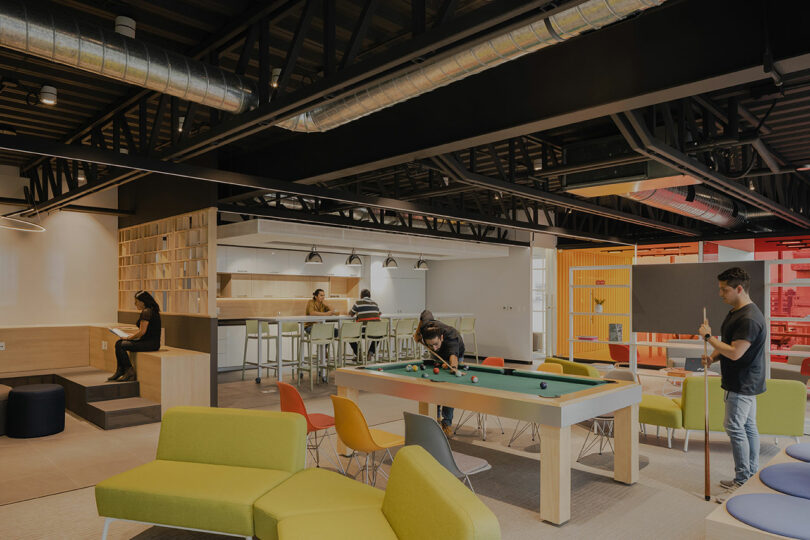 Modern office lounge with people sitting and playing pool. Features colorful seating, high-top tables, and industrial ceiling design.