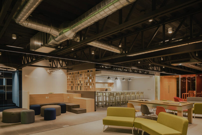 Modern office space with exposed ductwork, wooden partitions, colorful seating, and a bar area with stools.