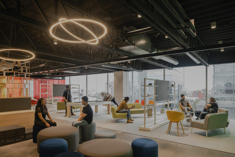 Modern open office space with people interacting, sitting on colorful furniture, and a city view through large windows.