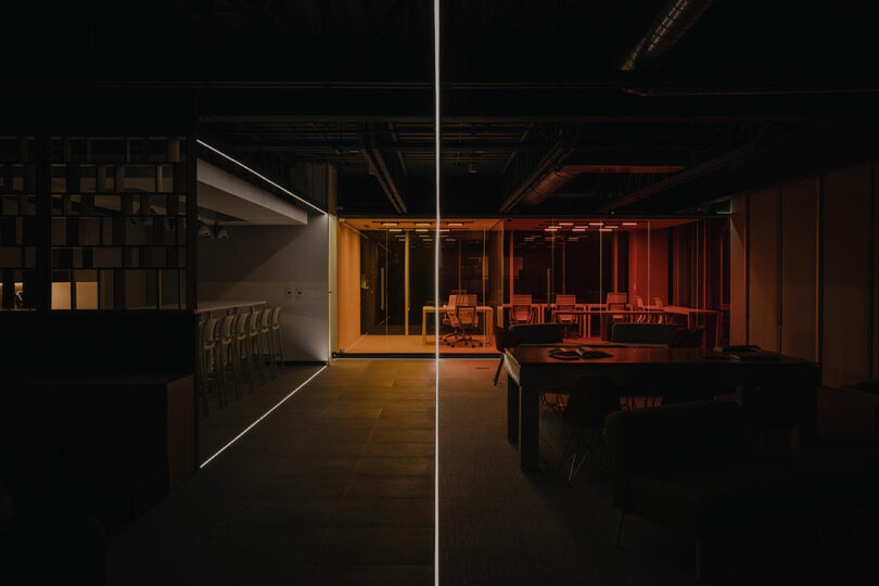 Dimly lit modern office space with a linear division. One side features a bar area illuminated in white, and the other a meeting area with red lighting. Minimalist furniture throughout.