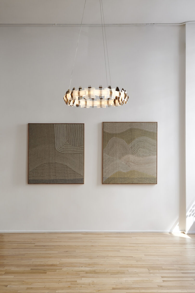 A modern chandelier hanging above two abstract paintings on a white wall with a wooden floor.