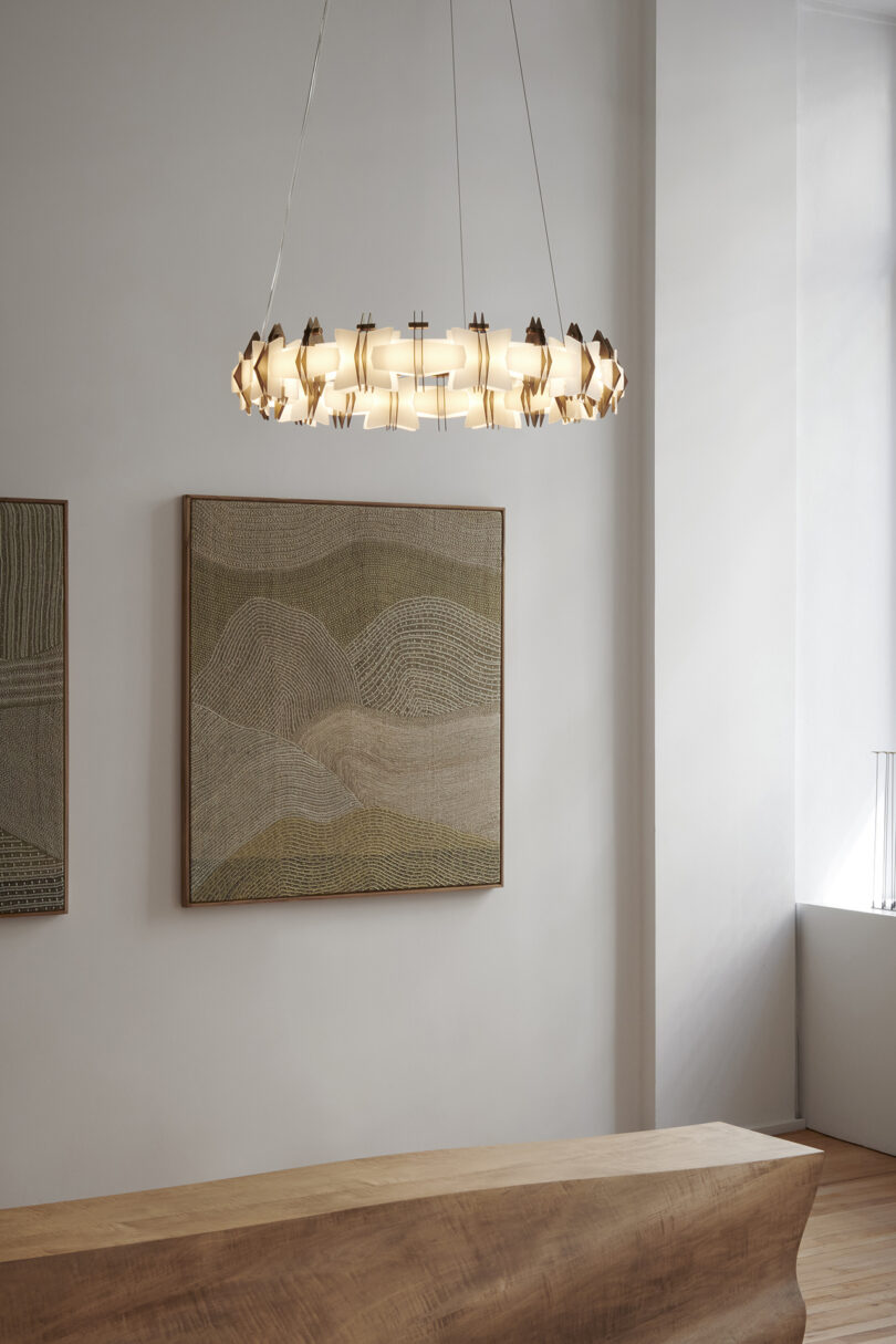 A modern chandelier hangs above a wooden surface. Two abstract textured paintings are mounted on the wall. Natural light illuminates the minimalist room.