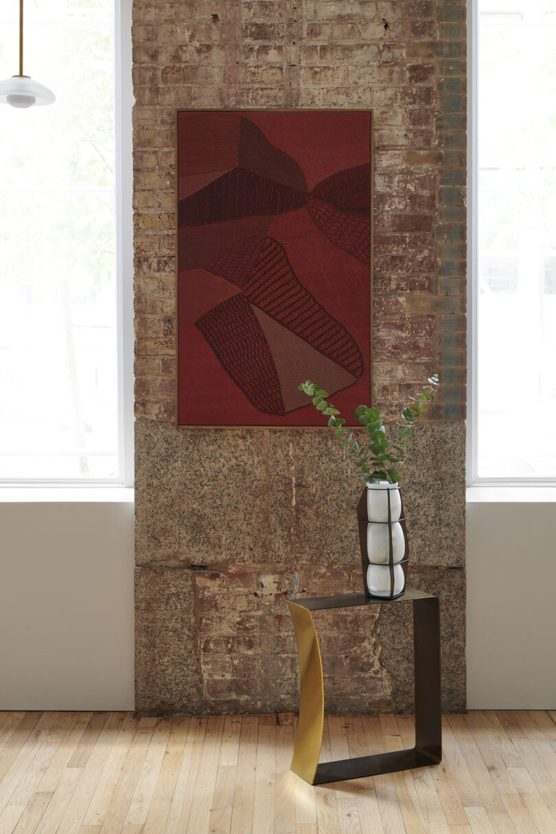 Wall art with abstract design on exposed brick wall; modern table holding a geometric vase with green foliage. Wood flooring and large windows in the background.