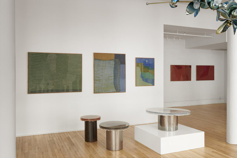 Art gallery interior with abstract paintings on white walls and modern round tables with metal bases on a wooden floor.