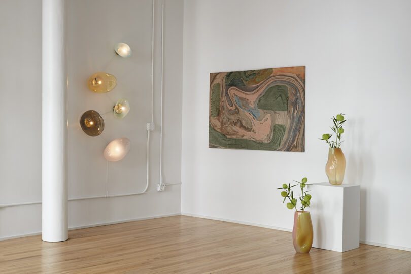 Art gallery corner with abstract wall painting, four oval wall lights, and two vases with plants on wooden floor.