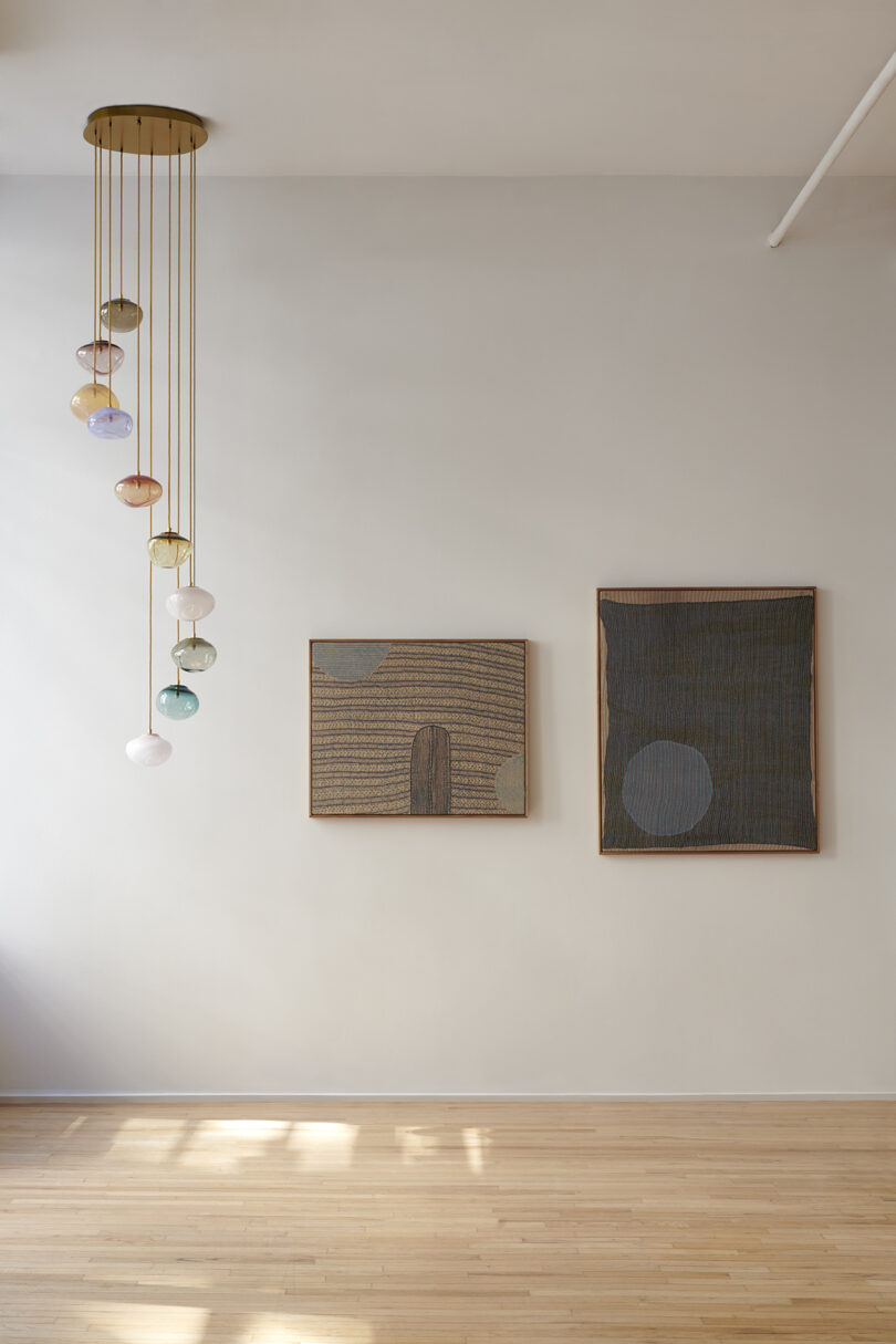 A minimalist room with two abstract paintings on a white wall and a modern hanging light fixture.