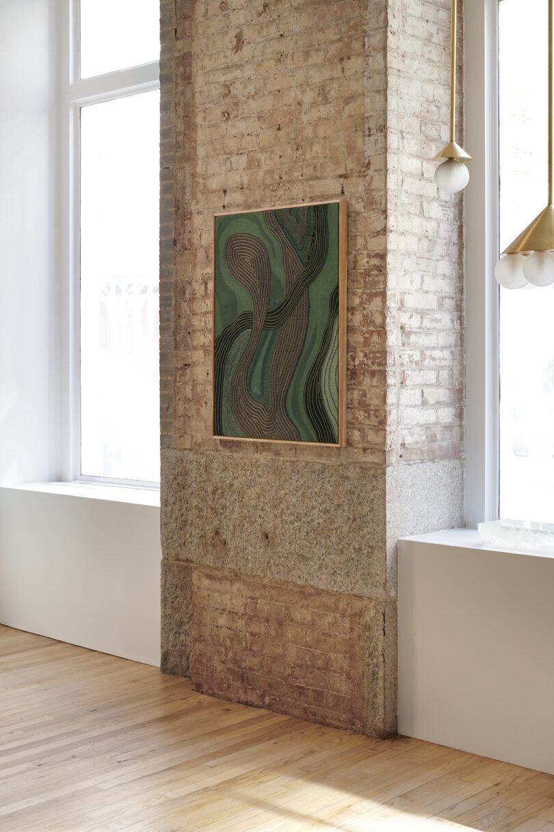 Embroidered artwork with abstract swirling patterns in greens and browns on a brick wall in a modern room with large windows and wooden floor.