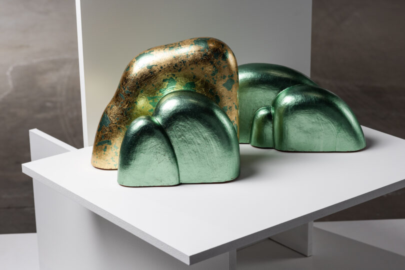 Two abstract green and gold sculptural forms rest on a white display stand in a gallery setting