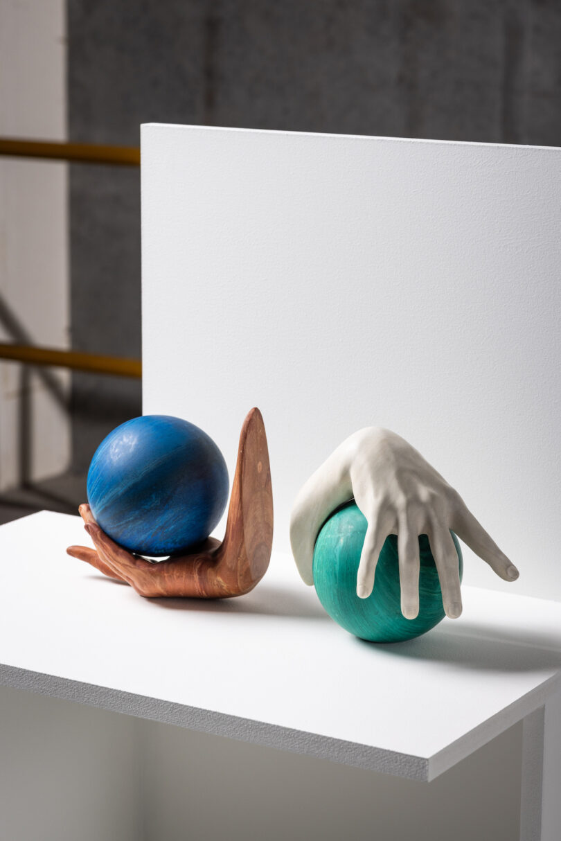 Two art sculptures on a white surface: a blue sphere resting on a wooden support resembling a hand, and a green sphere partially covered by a ceramic hand