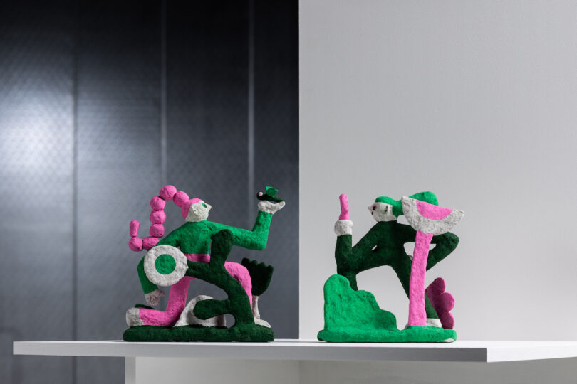 Two abstract sculptures on a shelf, featuring green and pink humanoid figures in dynamic poses against a grey background.