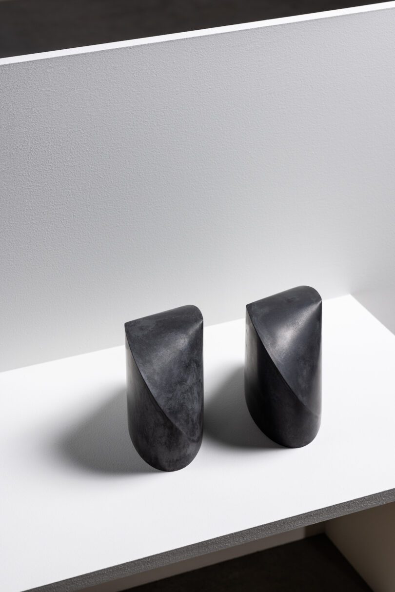 Two black, curved, cylindrical sculptures on a white surface with a white backdrop.