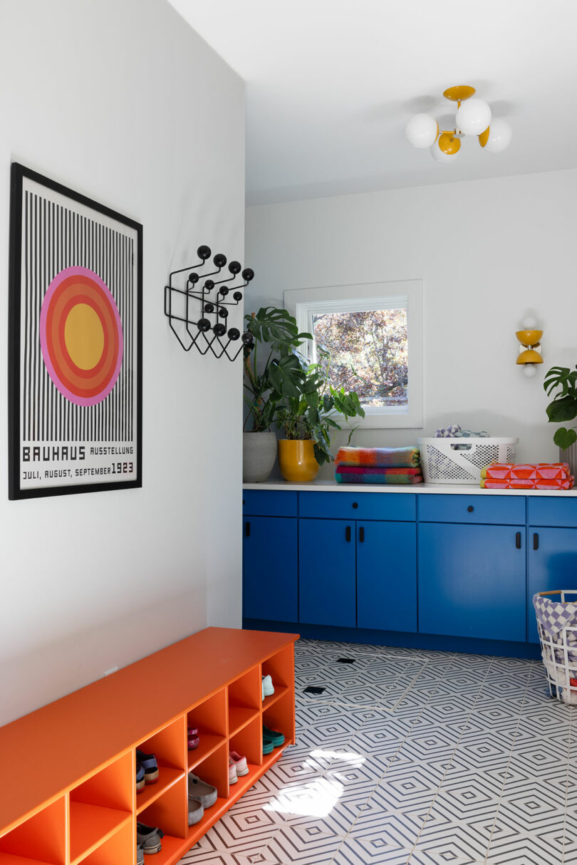 Room with geometric floor tiles, blue cabinets, an orange bench, and colorful decor. Wall art includes a Bauhaus poster. A small window and indoor plants are present.