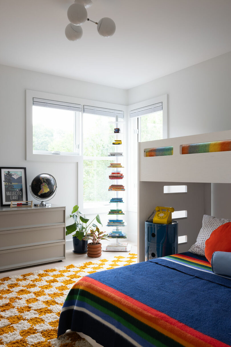 A bright bedroom with bunk beds, a colorful striped blanket, and a tall shelf of stacked books by the window. A globe and framed poster sit on a dresser. A yellow phone is on a nightstand.