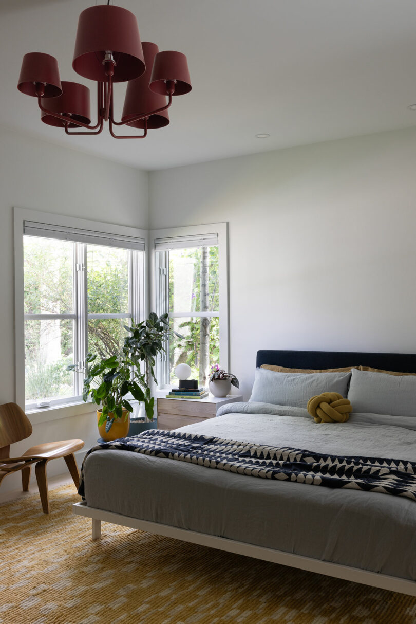 A bedroom with a bed covered in a geometric-patterned blanket, a wooden chair, a potted plant, and a window with greenery outside. A red chandelier hangs from the ceiling.