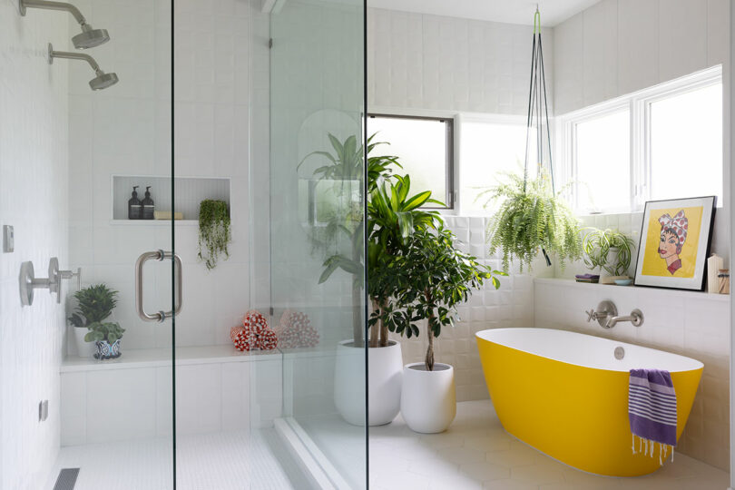 A modern bathroom with white tiles features a glass shower, a yellow bathtub, and several potted plants. A framed artwork is displayed above the tub.