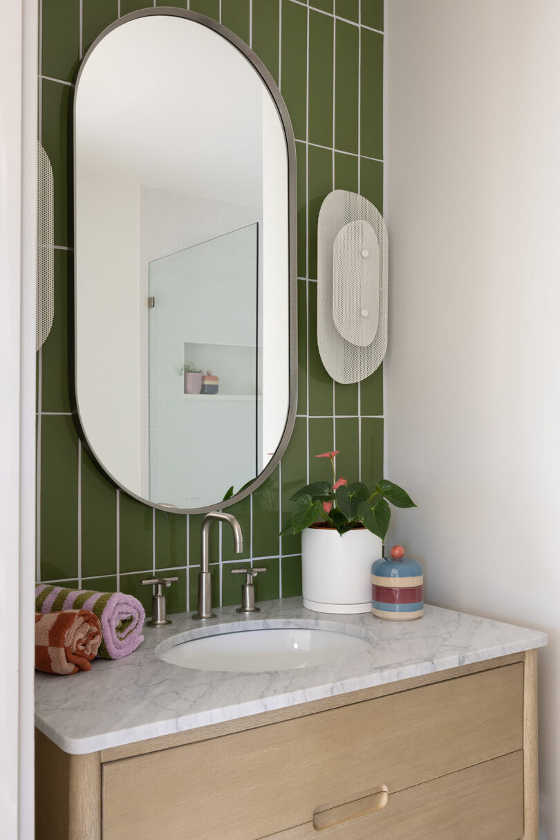 Bathroom with an oval mirror, green tiled backsplash, marble countertop, beige vanity, potted plant, and wall light. Towels and a container are on the counter.