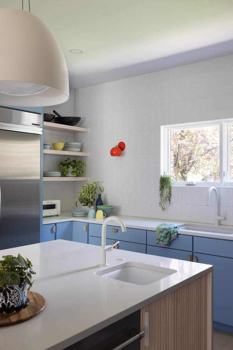 Modern kitchen with blue cabinets, white countertops, and a white island sink. Open shelves display dishes. Window above main sink, red wall lights, and potted plants as decor.