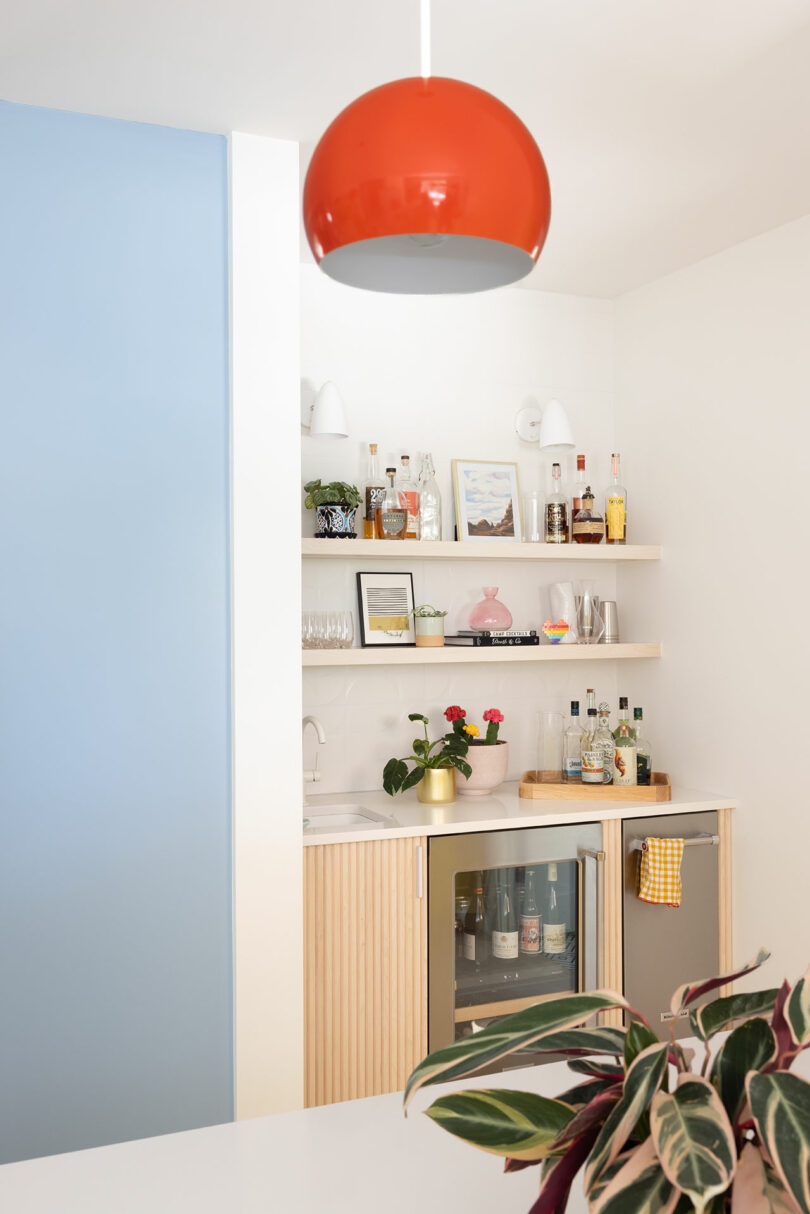 A modern home bar with wooden shelves displaying bottles and glasses, a small fridge, a blue accent wall, and a red pendant light.