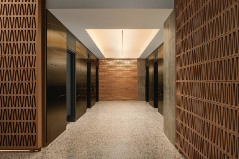 A modern hallway features three elevator doors with sleek, dark finishes and walls adorned with patterned wooden panels. The floor is tiled, and recessed lighting illuminates the space.