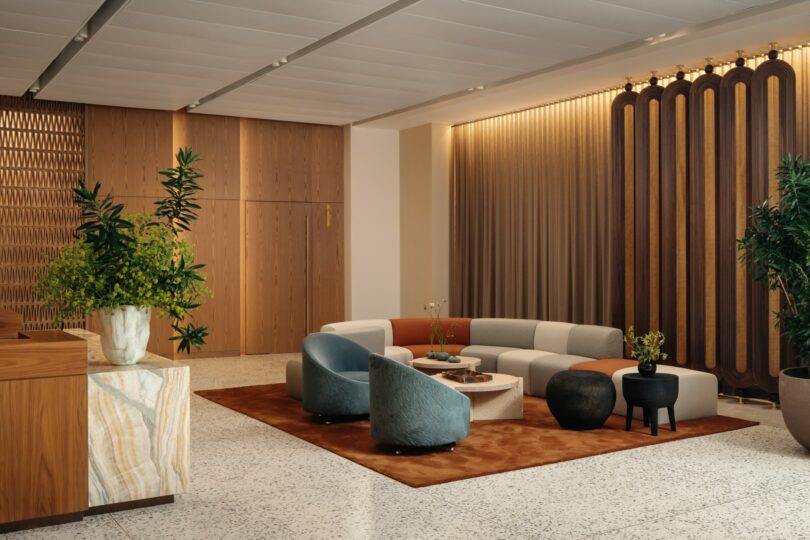 Modern lounge area with contemporary furniture including a curved sofa, chairs, and coffee tables on a brown rug. The room features wooden paneling, potted plants, and subdued lighting.