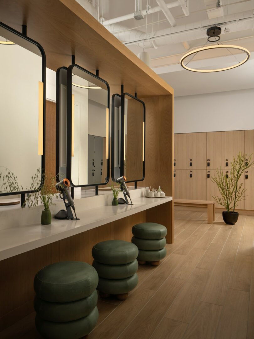 A modern, well-lit locker room features a vanity area with three green cushioned stools, large mirrors, hairdryers, and two plants. Light wood lockers are visible in the background.